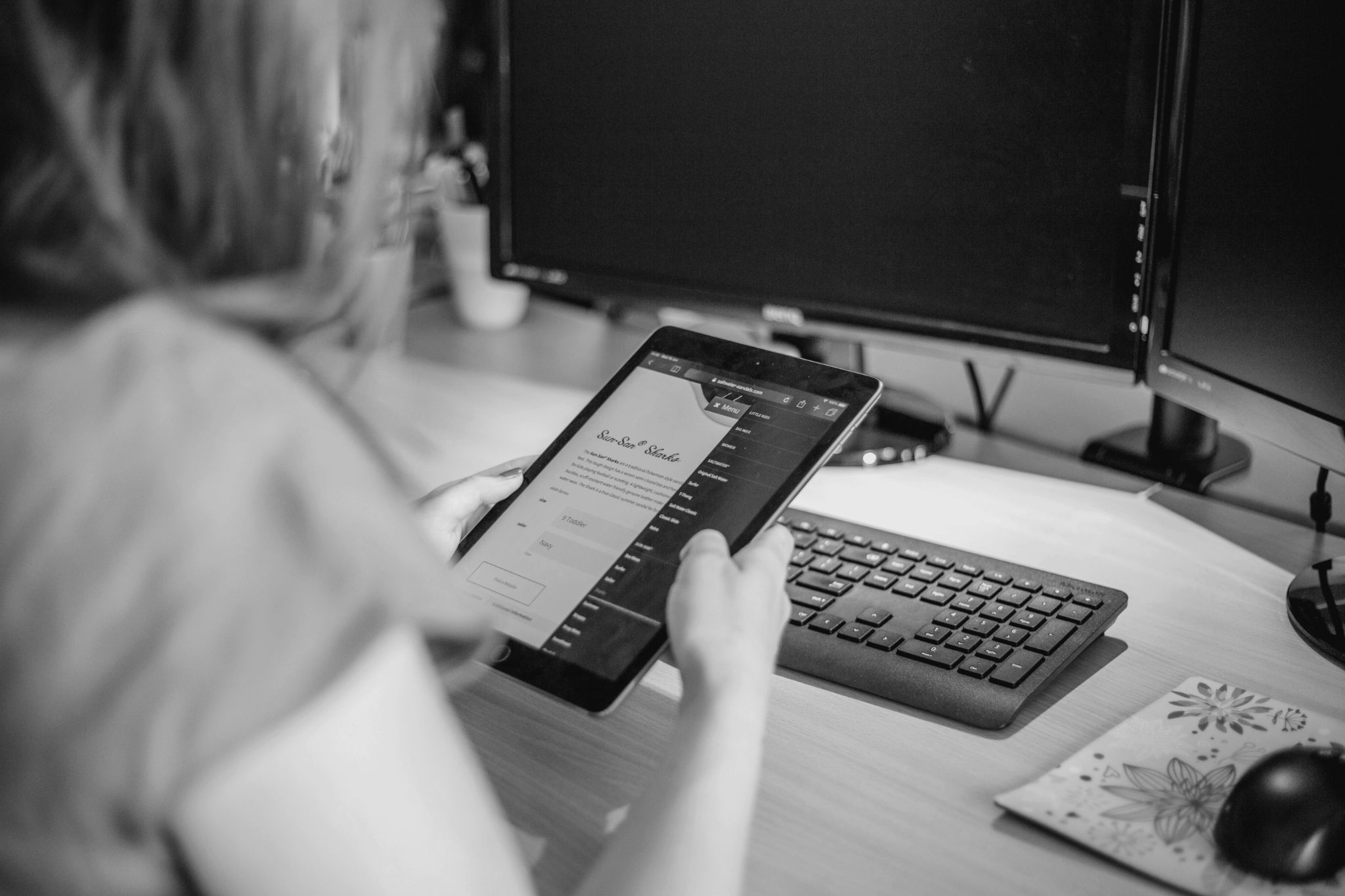 A person holding a tablet with a form visible on the screen, seated at a desk equipped with a computer keyboard and dual monitors, illustrating a modern workspace setup.